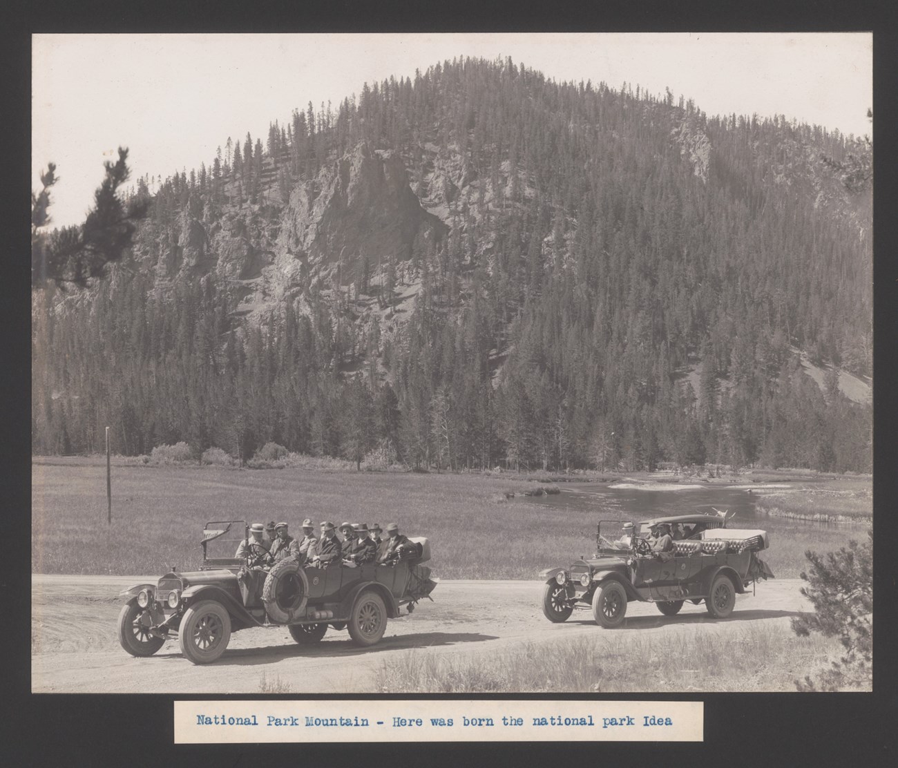 Men in two old open-top touring cars in front of a mountain