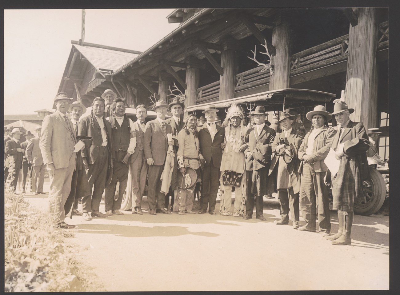 Men and suits and Native Americans pose for a photo in front of a large log building