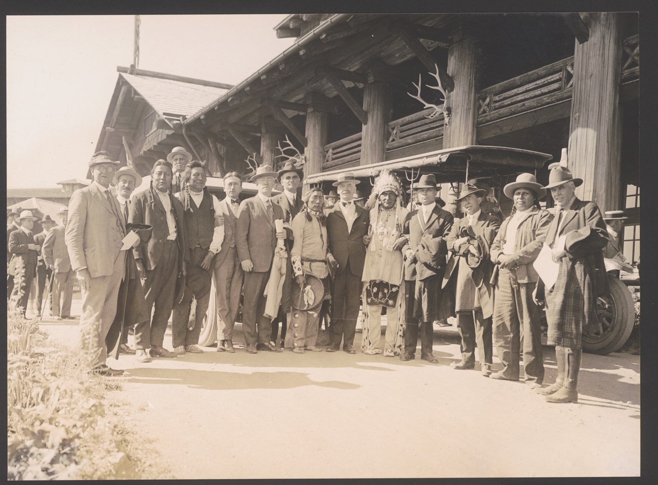 Delegation at Glacier Park Hotel Men and suits and Native Americans pose for a photo in front of a large log building