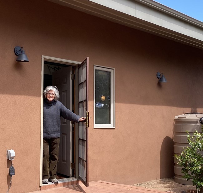 A woman with gray hair stands in the exterior doorway of an adobe home.
