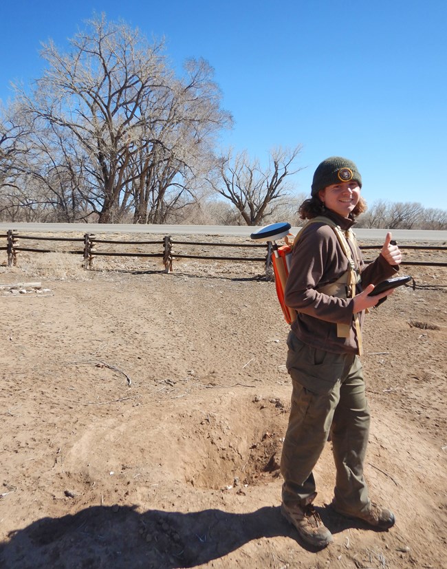 Shea giving a thumbs up as they stand beside a mound of dry earth with a hole in the center. They carry a tablet in their other hand and wear a neon orange backpack with a GIS device attached to the side.