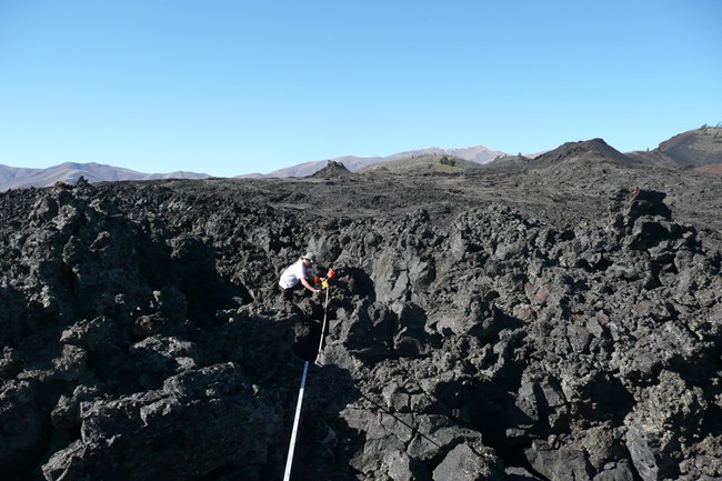 a biologist holding one end of a tape measure while standing on very rugged black rocks with cinder cones and mountains in the distance