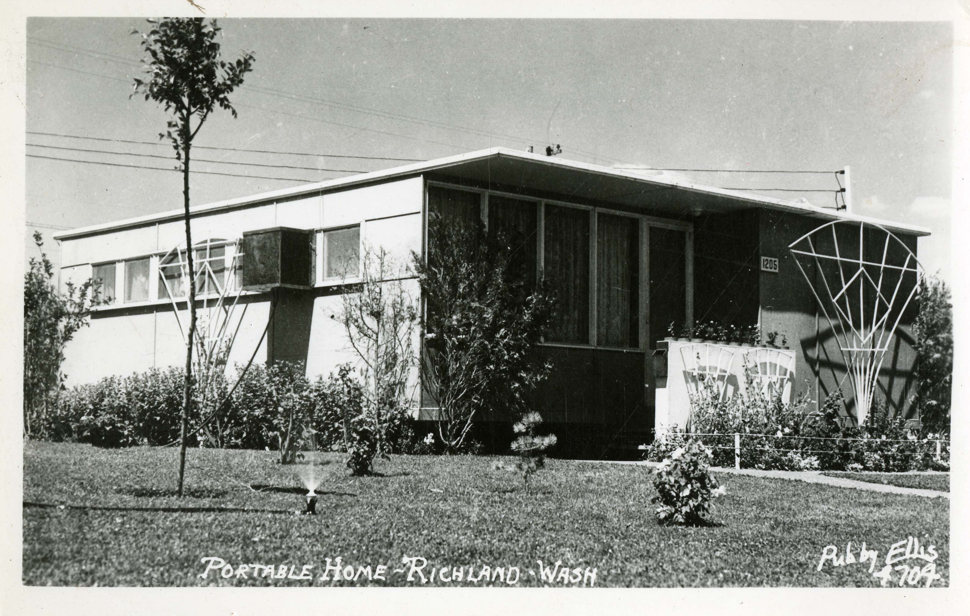 Square flattop house, shrubs, sprinkler in yard. Text: Portable home-Richland, WA Ruby Ellis 4704.