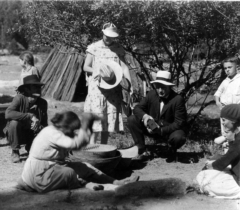 Ta-bu-ce demonstrates pounding acorns to a group of people standing and kneeling around her.