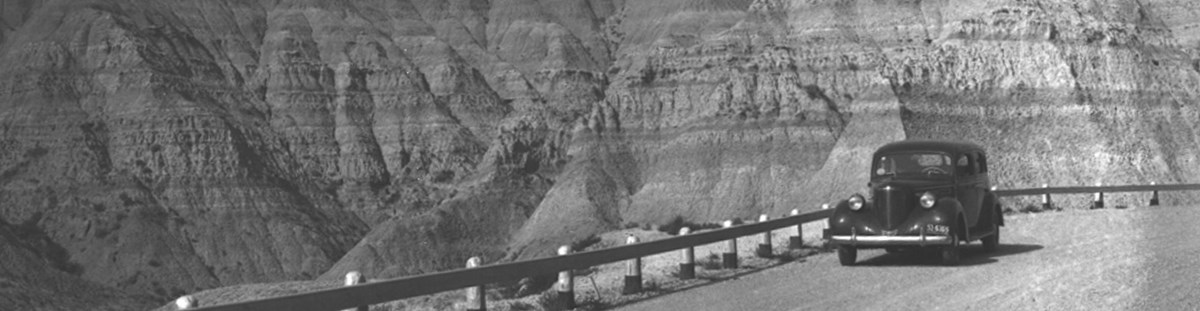 a black and white historic photo showing an old fashioned car driving in front of badlands formations.
