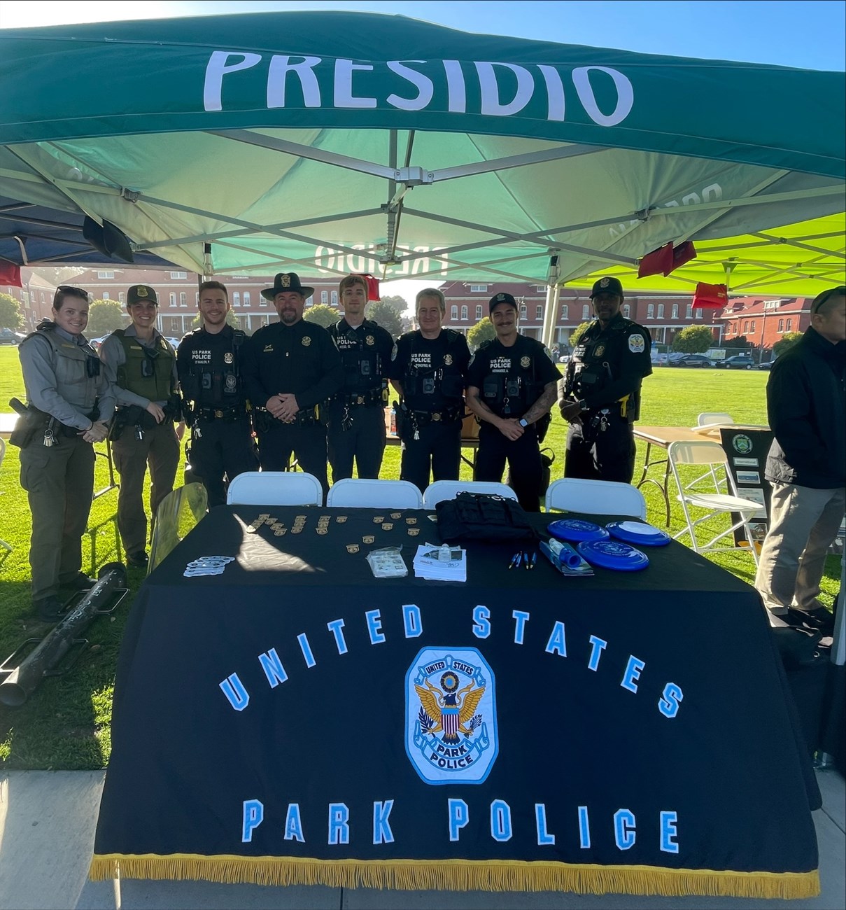 U.S. Park Police Table at National Night Out Eight U.S. Park Police officers stand behind a table with a blue tablecloth that says "United States Park Police" and has various giveaway items like frisbees and plastic police badges on it. The officers and table are all under a green portable canopy.