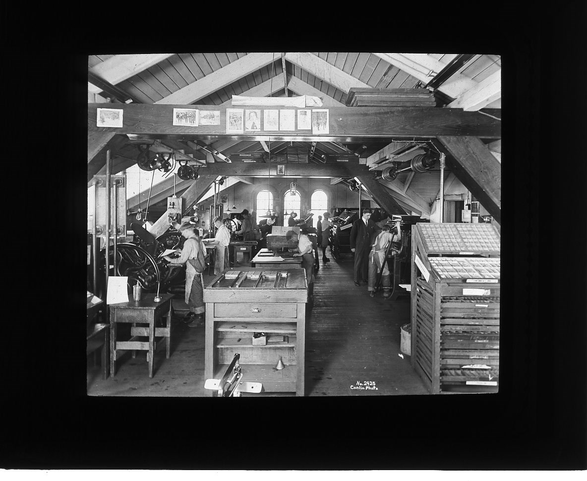 Boys work around a print shop, operating machinery while one teacher helps a student. There are multiple tables in the center of the room. A beam by the ceiling is lined with some papers with prints on them.