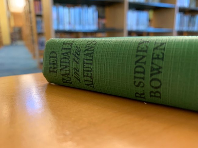 Photo of the spine of a green book on a table in the library. Text: Red Randall in the Aleutians. R. Sidney Bowen.