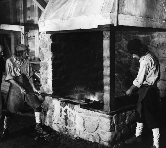 Two men in white shirts and dark hats standing around a fire.