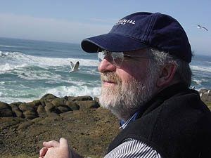 Robert Melnick sitting beside a rocky seashore. Robert Melnick in a cap sitting beside a rocky seashore looking out to sea with gulls in the sky.
