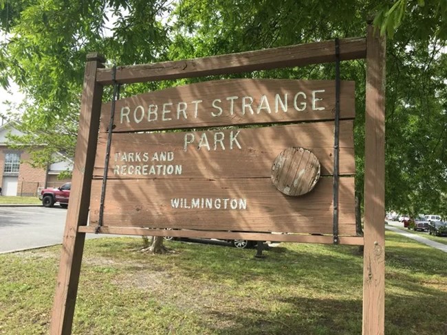 A wooden sign with text "Robert Strange Park: Parks and Recreation, Wilmington" in front of grass and trees