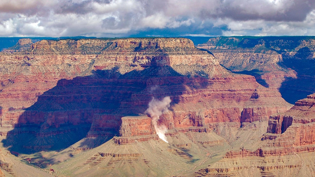 Vermilion colored stratified cliffs in a canyon wall. A plume of dust is visible from a rockfall in progress. Large summer clouds shade portions of the landscape.