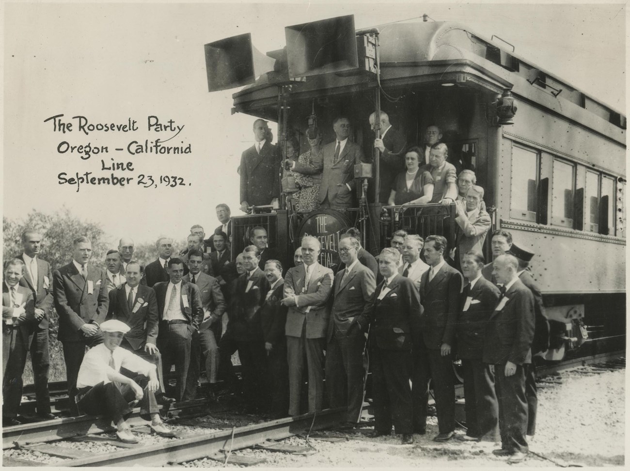 Group of men stand at the back of a train car. FDR is at the back railing. JPK stands below on the train tracks.