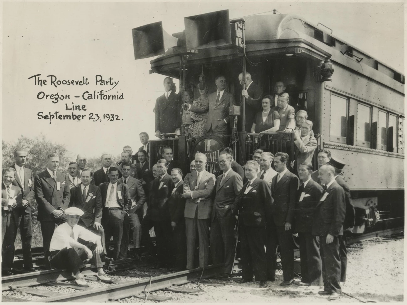 Group of men stand at the back of a train car. FDR is at the back railing. JPK stands below on the train tracks.