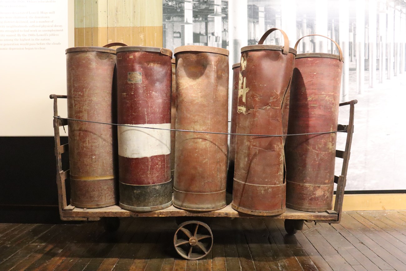 roving can_1 A wooden cart holding two rows of five large metal tubes. The tubes are a red brown color and some are slightly crushed