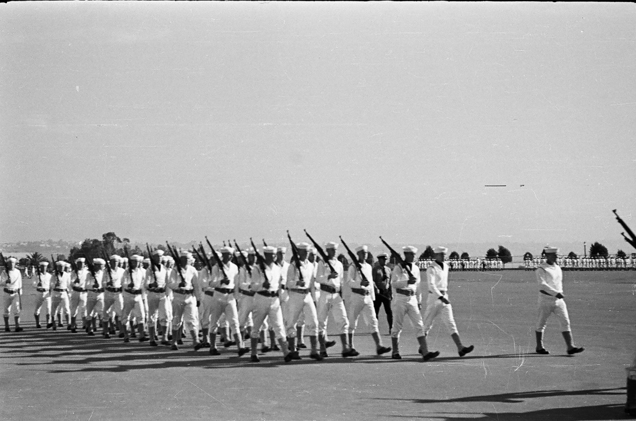 men in white uniforms with guns over their shoulders marching across a field in unison.
