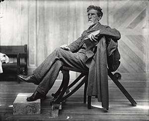 saint-gaudens man with dark hair and white beard sitting in a chair with his feet propped up on a stone slab