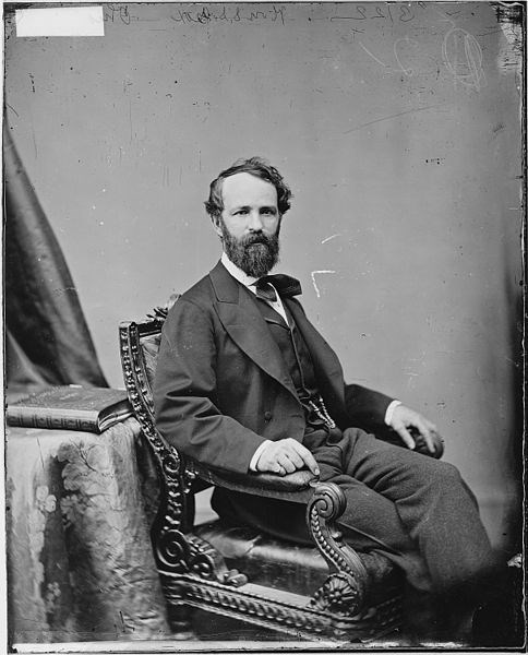 man in asuit has a thick brown beard. he is sitting in a chair with both hands holding the arms
