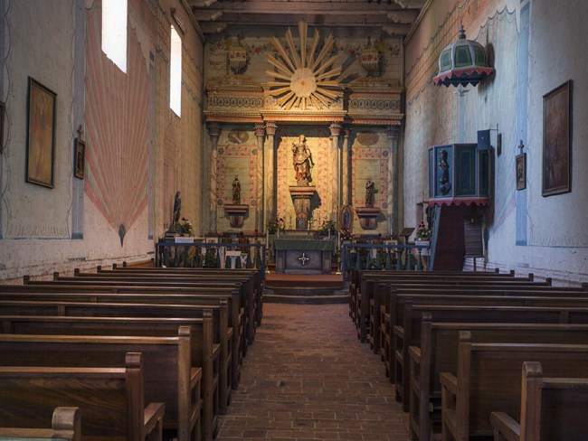 Interior of Mission, facing a colorfully painted altar with wooden pews on either side of center aisle