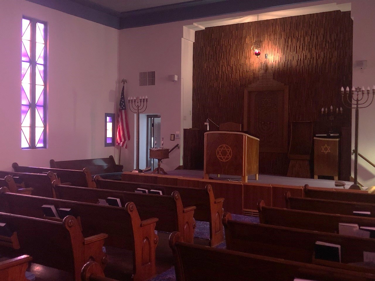 B'nai Israel Synagogue sanctuary Interior of a synagogue illuminated by a stained glass window. The bimah includes a centered reading table, a suspended lamp, and a textured wall.