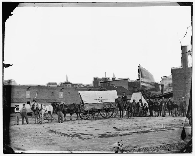 Black and white photograph of two horses pulling a wagon titled U.S. Sanitary Commission with a large group of people at the far end of the wagon. There are brick buildings in the background.