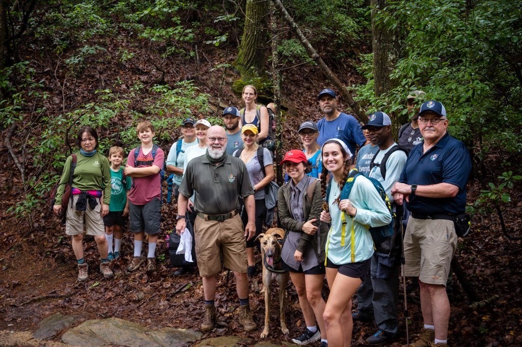 Group of 17 people of varied ages stand smiling looking forward in forest setting, dressed in hiking clothes