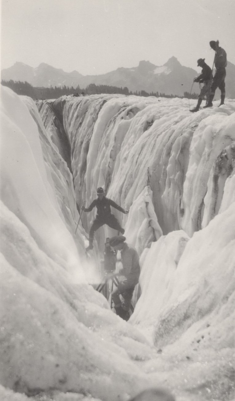 Man straddles a crevasse in front of a movie camera as several men look on.