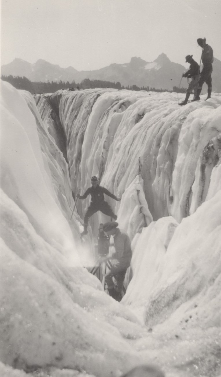 Filming the crossing of a crevasse. Man straddles a crevasse in front of a movie camera as several men look on.