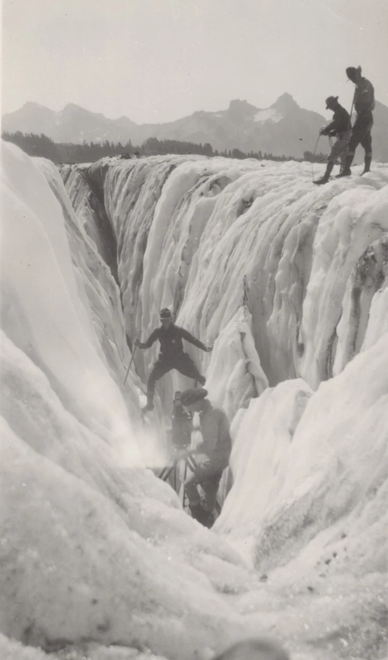 Filming the crossing of a crevasse. Man straddles a crevasse in front of a movie camera as several men look on.