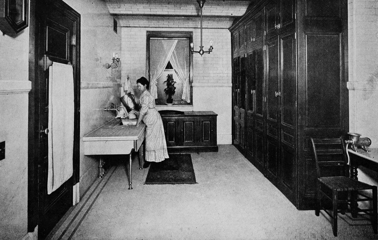 lt= A woman washing dishes in a large scullery.