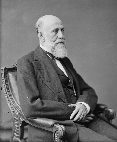 older gentleman has a long white beard and is sitting in a chair with hands folded on  lap