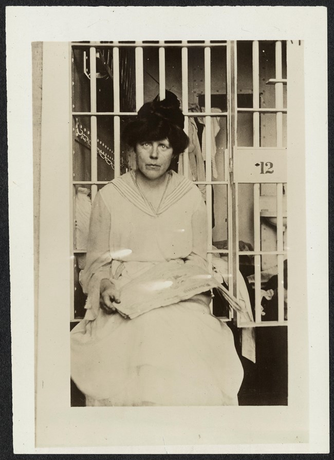 Lucy Burns in her cell at the Occoquan Workhouse, Lorton, VA. Informal portrait, Lucy Burns, three-quarter length, seated, facing forward, holding a newspaper in her lap in front of a prison cell, likely at Occoquan Workhouse in Virginia.