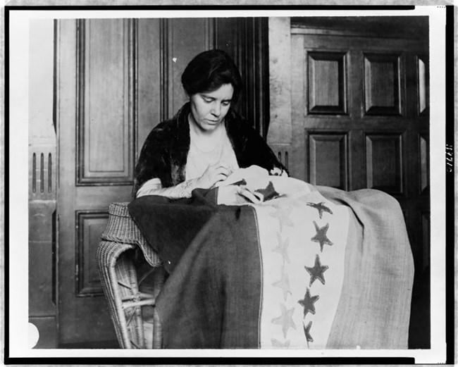 Alice Paul sewing a suffrage flag. Alice Paul, half-length portrait, seated, facing slightly right, sewing suffrage flag.