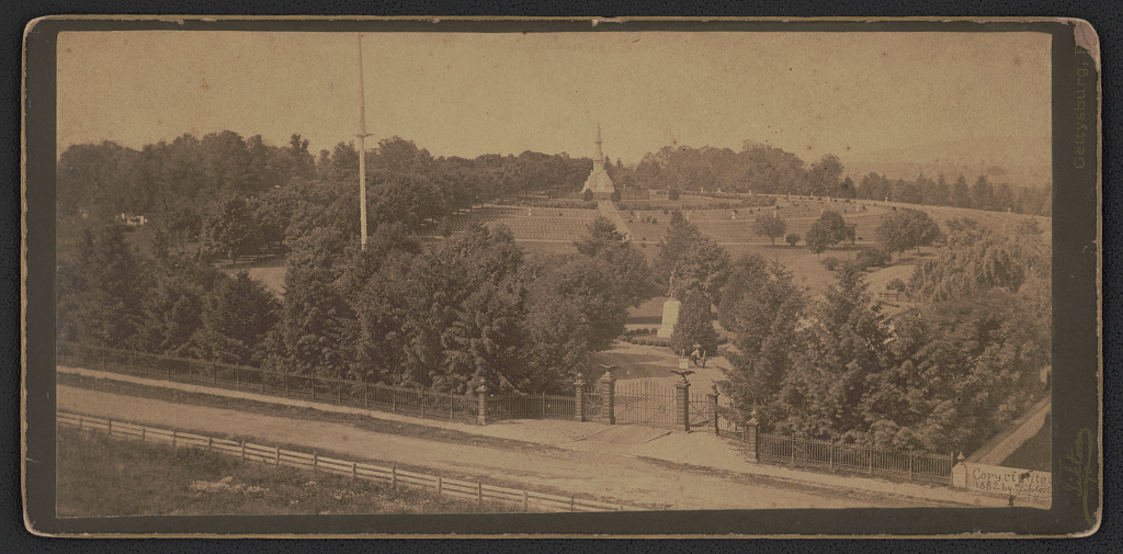 Photograph shows gate to Gettysburg National Cemetery in front of statue of Major General John F. Reynolds with the Soldiers' National Monument in the distance, surrounded by grave markers.