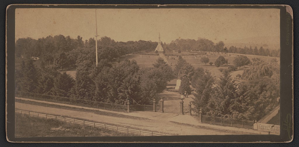 Photograph shows gate to Gettysburg National Cemetery in front of statue of Major General John F. Reynolds with the Soldiers' National Monument in the distance, surrounded by grave markers.