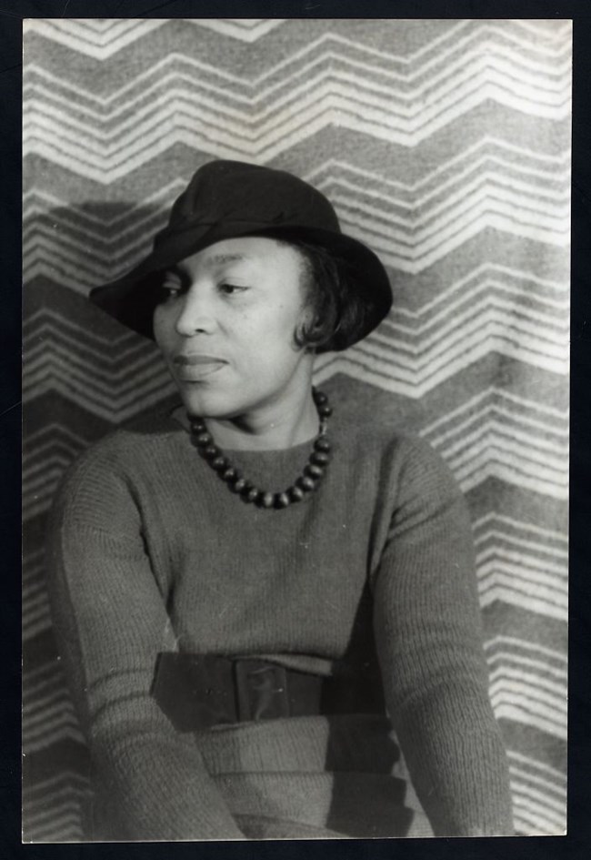 Zora Neale Hurston A Black woman sits looking to the left wearing a hat, beaded necklace, and sweater.