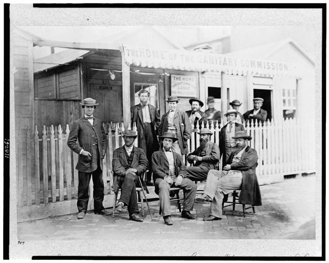 Black and white photograph of group of men, some seated some standing, all wearing hats and dark suits stand in front of wood building called the home of the sanitary commission