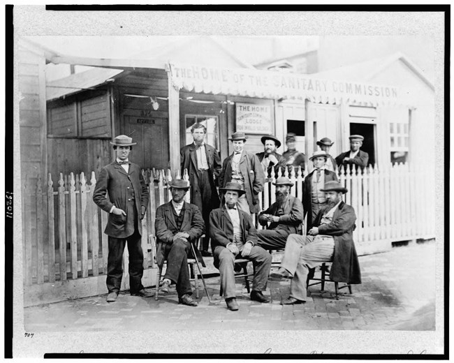 serviceusc Black and white photograph of group of men, some seated some standing, all wearing hats and dark suits stand in front of wood building called the home of the sanitary commission