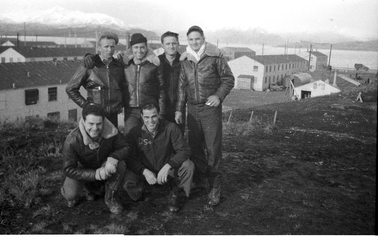 Group of 6 men posing on hill in front of two story white  buildings, the ocean, and snowy mountains.