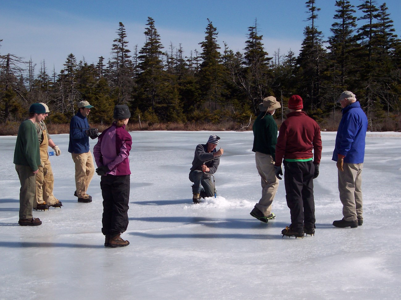 people stand around an individual using an auger to drill into ice