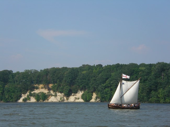 A replica of an English barge sails on a river with high cliffs for banks.