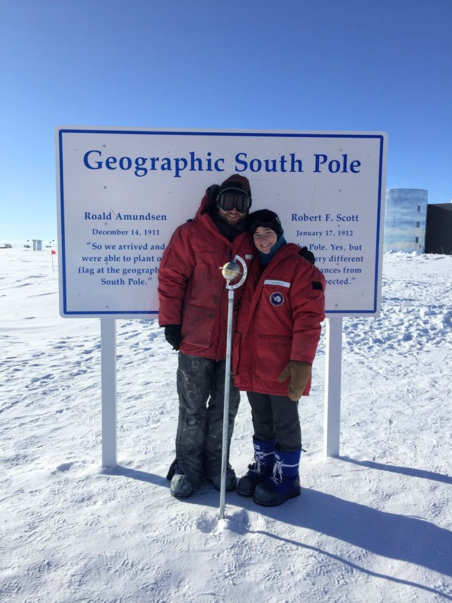 Two field biologist pose for photo wearing very large red parkas, warm boots, gloves, and hats at the Geographic South Pole.