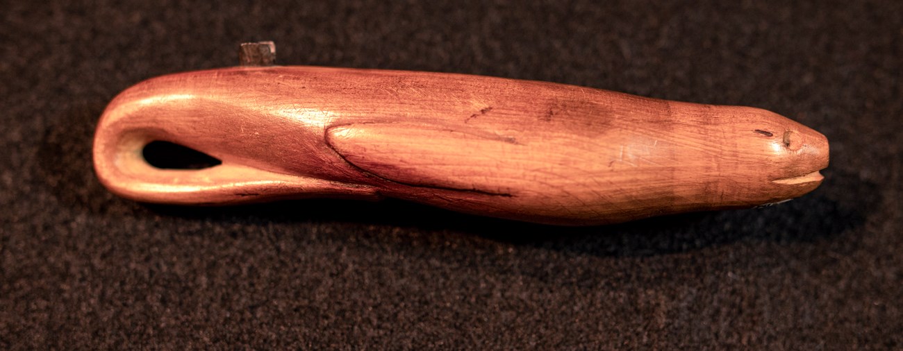 Wooden tool carved in a seal shape. A piece of metal on its back splits grasses for basketweaving.