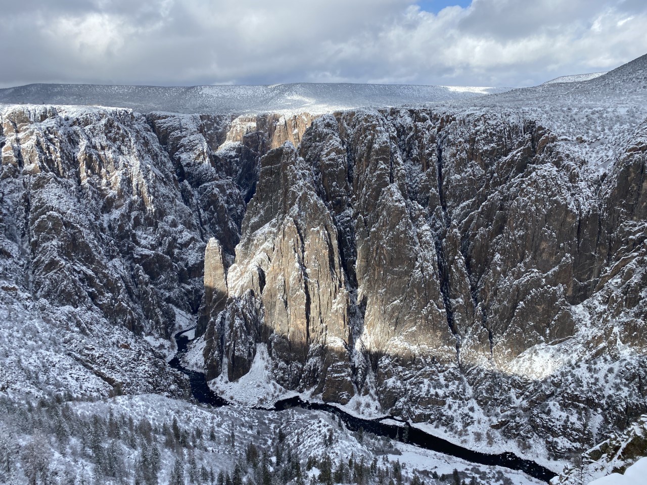 Black Canyon in winter Snow covering sections of steep dark brown canyon walls. A dark river is at the bottom of the canyon. Snow lingers on the canyon rim. Sky has clouds.