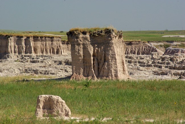 an elevated portion of prairie stands over green grasses and in front of badlands canyons.