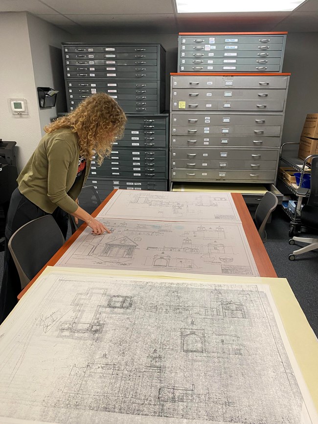 Woman with blonde hair points at a map on a large table with two other maps on it. A wall of two map cabinet stacks are behind her