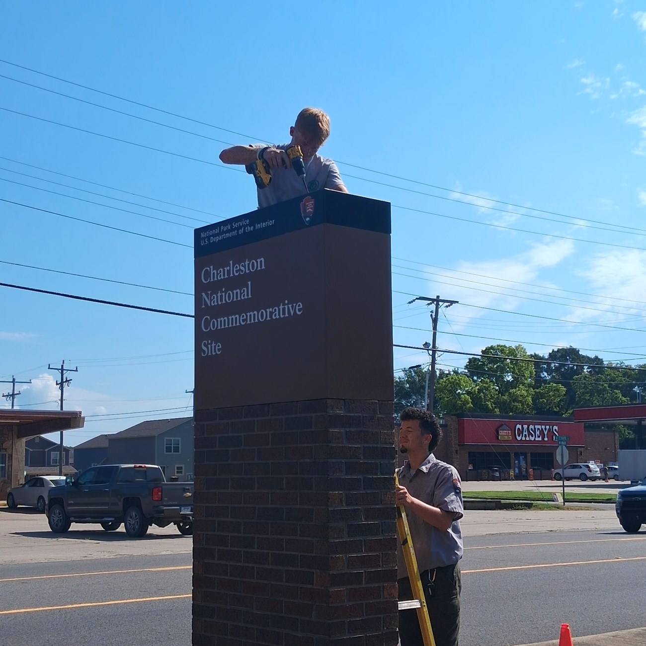 Two people install a Charleston National Commemorative Site sign, with one person drilling the sign on the top of a brick tower and the other holding the ladder steady.