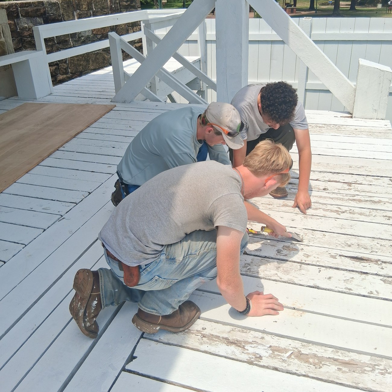 Three people kneel on a white wooden platform, scraping and repairing the weathered boards.