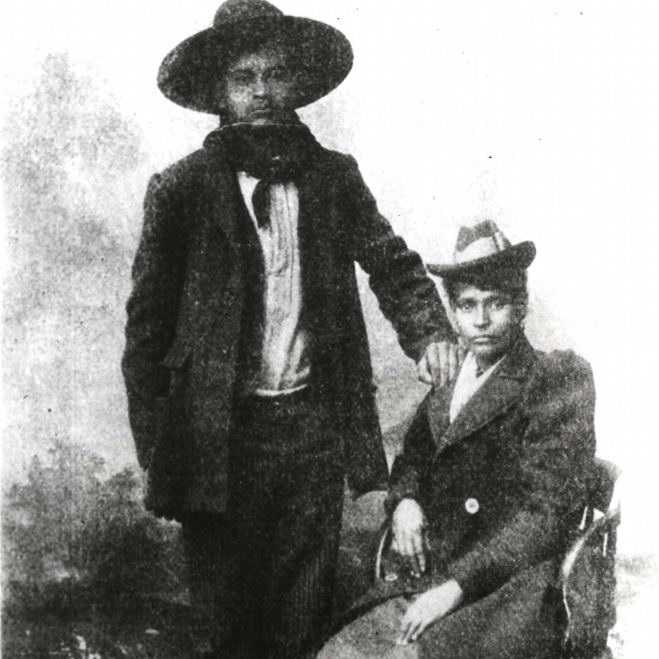 Crawford Goldsby alias Cherokee Bill and his mother Ellen Lynch Black and white historic image of Cherokee Bill standing wearing a dark wide-brim hat with his hand on the shoulder of the woman sitting next to him.