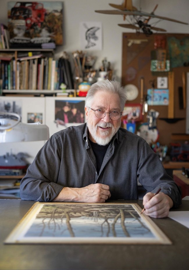 smiling man white short white hair, glasses, and a dark blue shirt sits in front of a drawing with a pencil in his hand